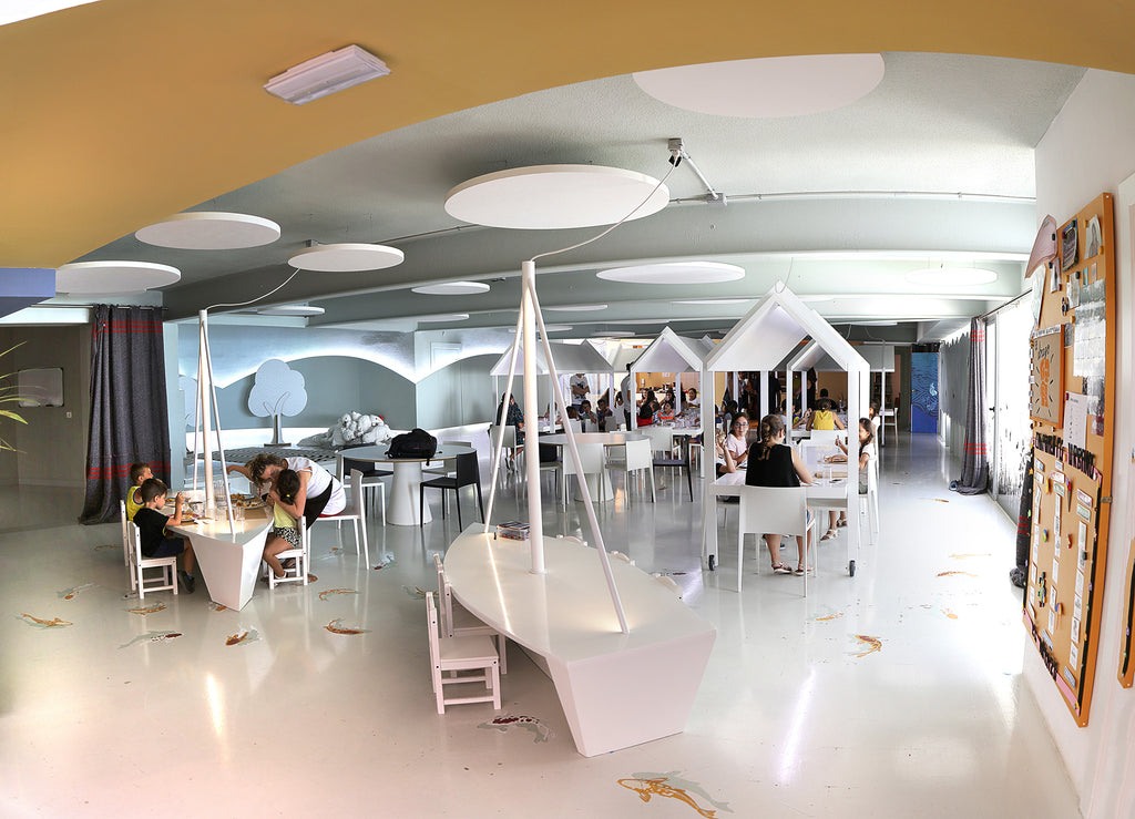Sala multifuncional que contiene mesas con formas de barcos o casas. Espacio  con mucha luz y tonos neutros.